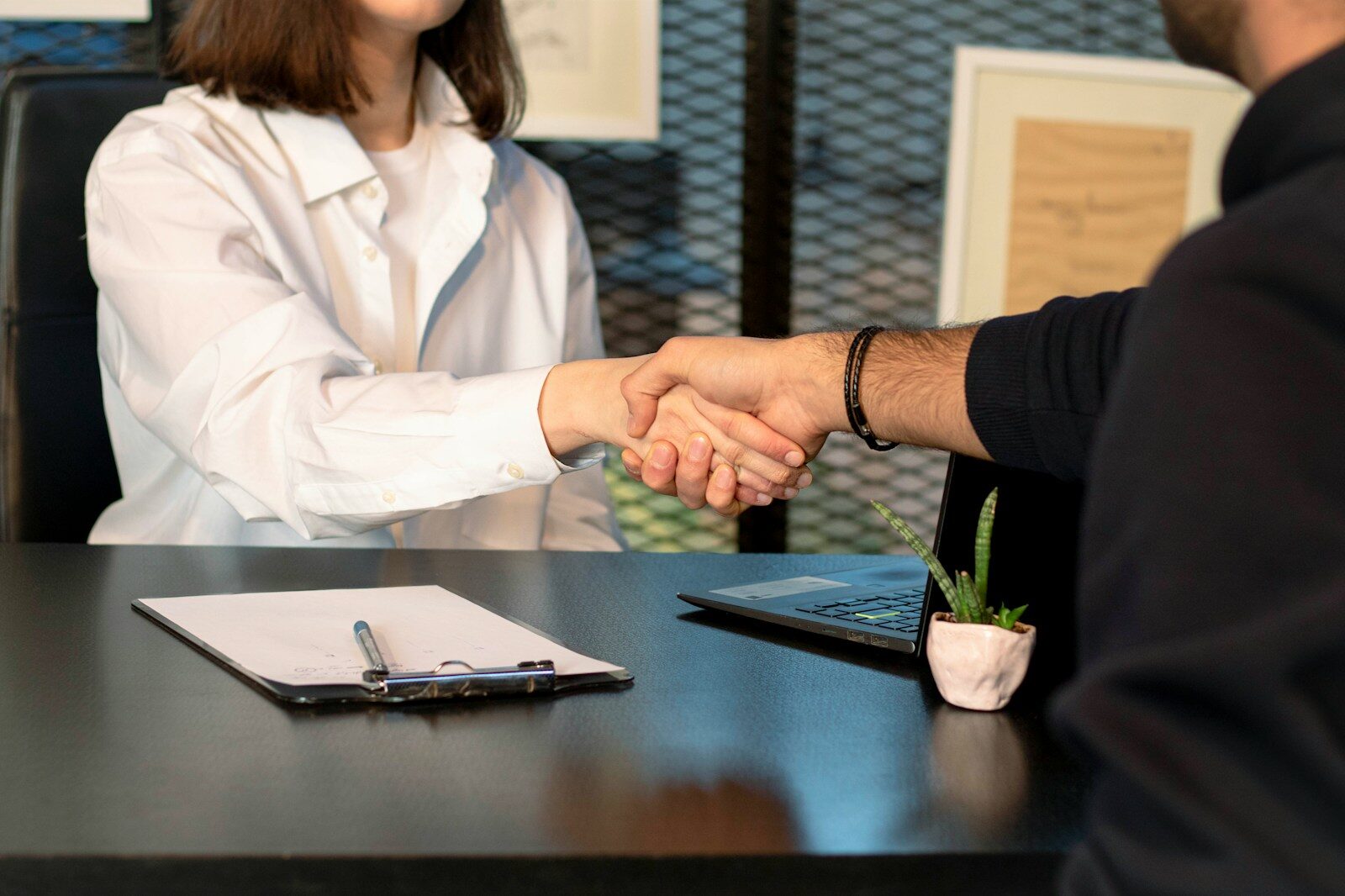 Professional handshake between two individuals at an office desk, symbolizing a successful agreement or partnership. Items on the desk include a clipboard and potted plant.