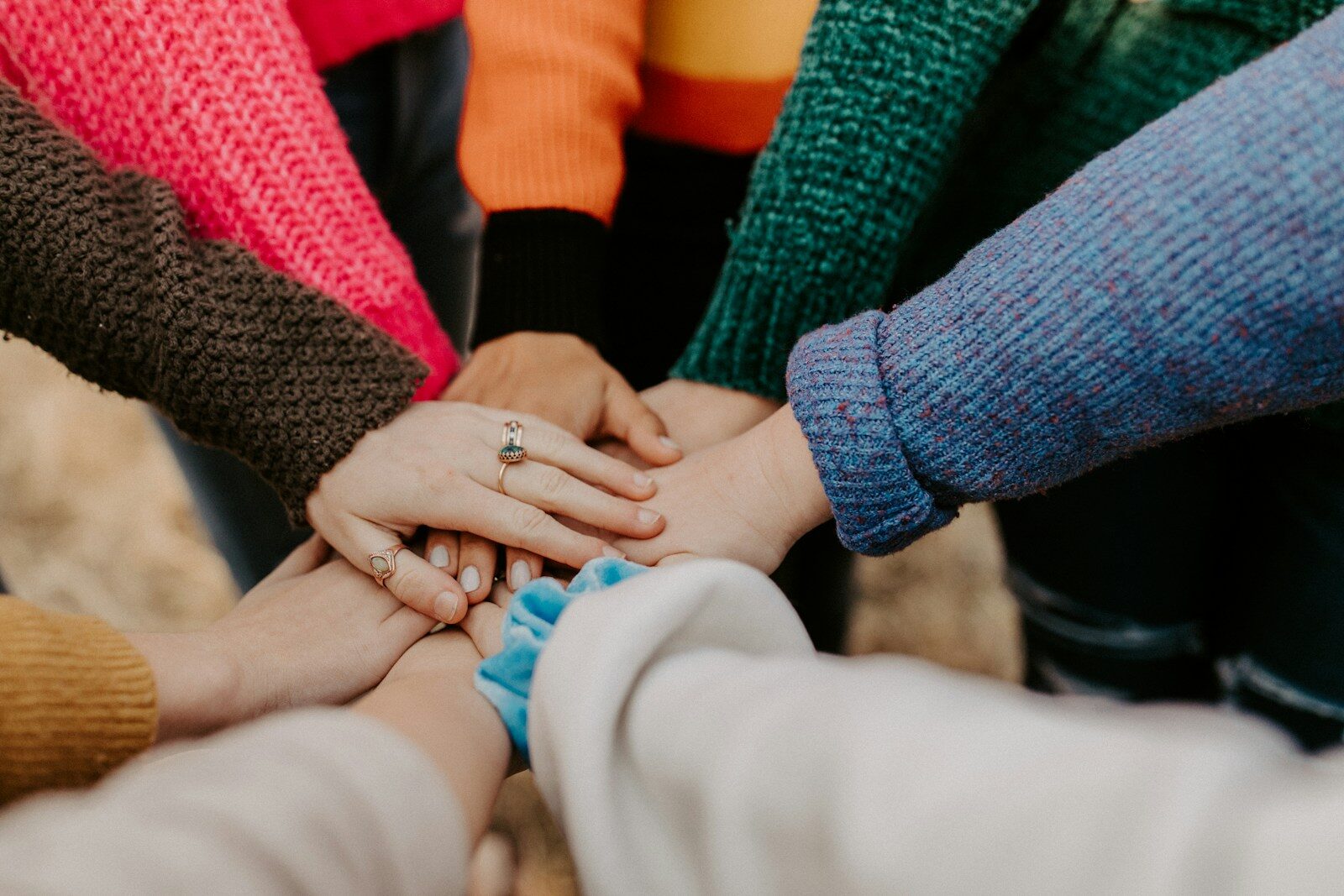 Hands of diverse individuals resting on one another, showcasing unity and connection, each adorned with colorful sweaters and accessories.